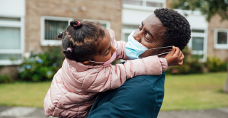 A father, wearing a face mask, hugs his young daughter